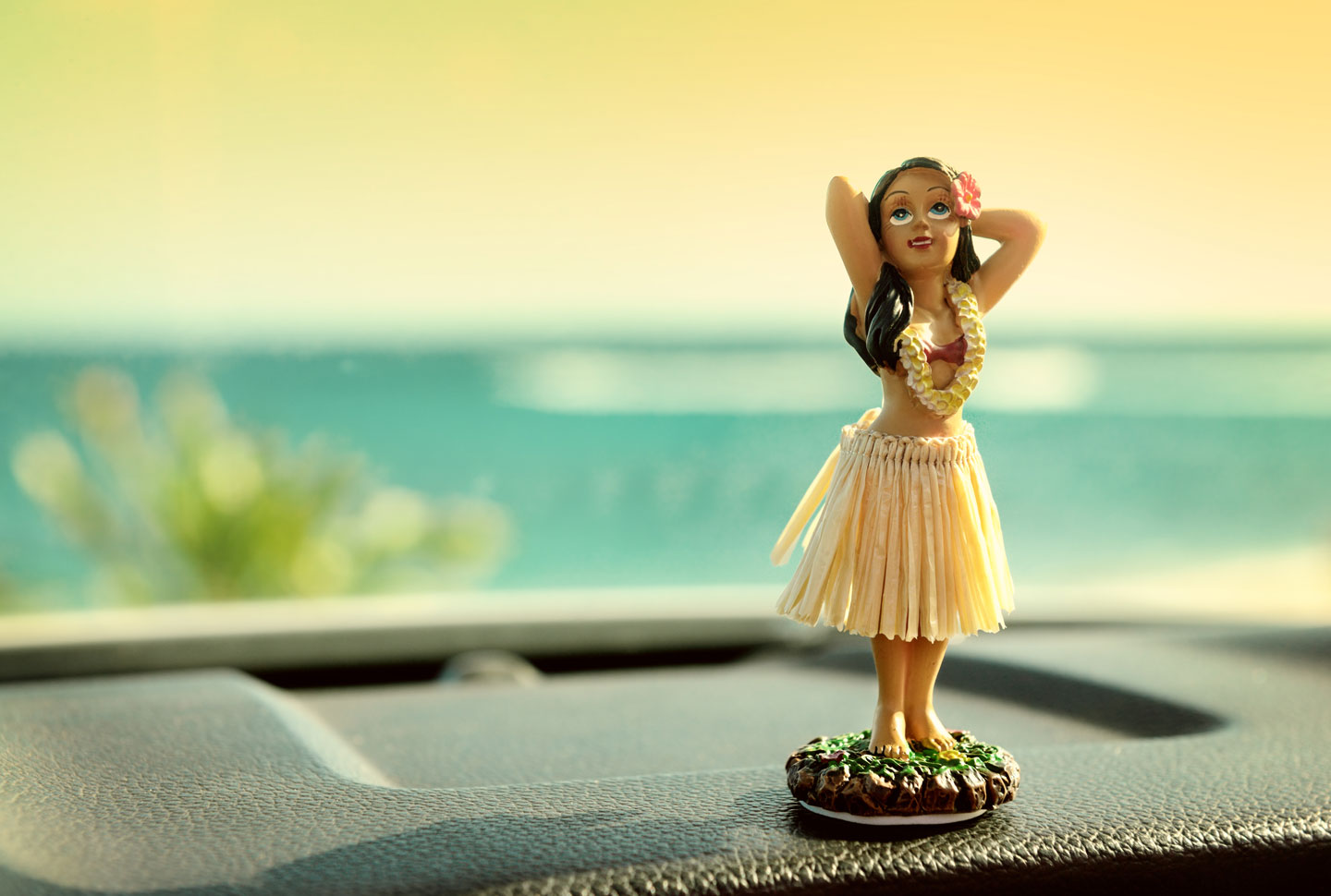 Hula-Figurine in Tanzpose auf einer Autokonsole, Hintergrund mit unscharfem Blick auf das Meer und blauem Himmel.